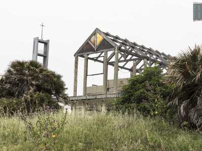Ora davanti a noi c'� un piccolo terreno incolto, mentre in lontananza svettano lo scheletro del tetto spiovente dell'hotel, senza muri e sorretto dalle sole colonne e il campanile della vicina parrocchia Stella Maris